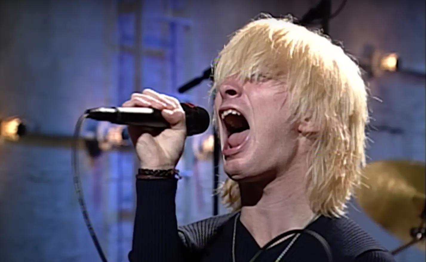 Thom Yorke passionately singing with mouth wide open, holding a microphone, wearing black sweater and necklace, with shaggy platinum blonde hair covering his eyes during a live Radiohead performance.